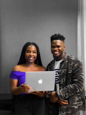 Two people smiling while standing close together, with one holding a laptop displaying an Apple logo. The person on the left is wearing a purple off-shoulder top, and the person on the right is wearing a camouflage jacket over a t-shirt.