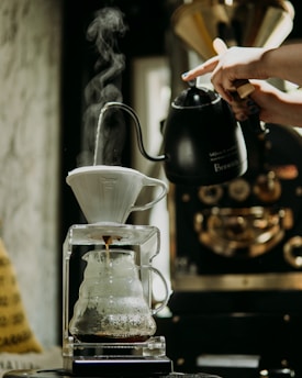 A hand pours hot water from a black kettle into a white coffee dripper. Steam rises from the dripper as fresh coffee drips into a glass server below. The background features a blurred coffee roasting machine.