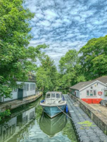 Aqua current boat docked beside a charming canal-side village with lush greenery.