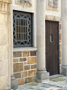 A section of a building facade features a stained glass window with geometric patterns and a dark wooden door with a small window. The facade is constructed with stone and brickwork, incorporating decorative details such as smooth columns and textured panels.