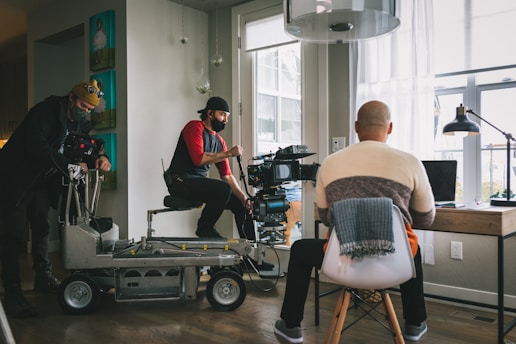 A film crew is setting up a scene in a room with large windows. Two crew members are operating a camera on a dolly while another person sits with their back to the camera at a desk with a laptop, appearing as if they are acting or being filmed.