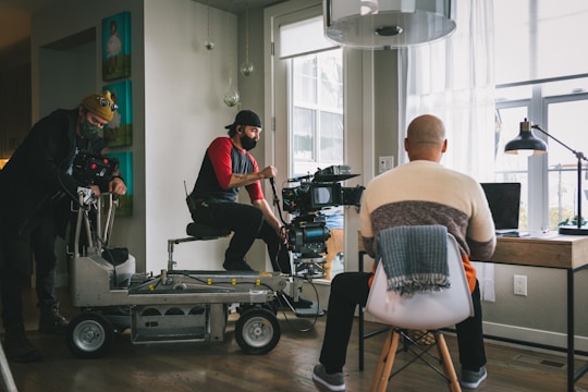 A film crew is setting up a scene in a room with large windows. Two crew members are operating a camera on a dolly while another person sits with their back to the camera at a desk with a laptop, appearing as if they are acting or being filmed.