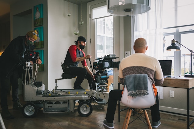 A film crew is setting up a scene in a room with large windows. Two crew members are operating a camera on a dolly while another person sits with their back to the camera at a desk with a laptop, appearing as if they are acting or being filmed.