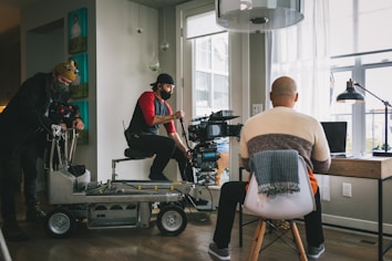 A film crew is setting up a scene in a room with large windows. Two crew members are operating a camera on a dolly while another person sits with their back to the camera at a desk with a laptop, appearing as if they are acting or being filmed.