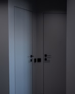 Two minimalist, modern doors with sleek black handles set in a dark corner, casting subtle shadows. The walls and doors have a smooth, matte finish, contributing to a clean, contemporary look.