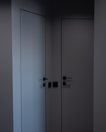 Two minimalist, modern doors with sleek black handles set in a dark corner, casting subtle shadows. The walls and doors have a smooth, matte finish, contributing to a clean, contemporary look.