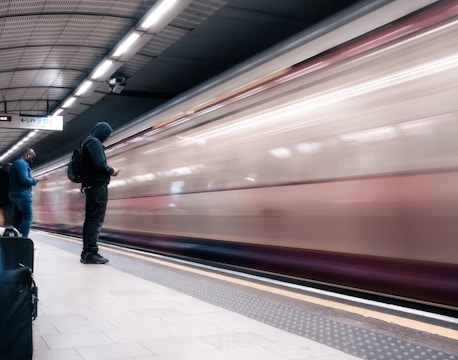 A fast-moving train blurs past on a subway platform where two individuals stand engrossed in their mobile devices. The scene includes a suitcase nearby on the platform floor.