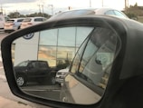 A reflection in a car side mirror shows a parking area with several vehicles, including a dark-colored truck and a light-colored SUV. The background features a building with large windows reflecting a sunset. The Volkswagen logo is visible on the building and on the vehicle registration in the mirror.