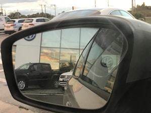 A reflection in a car side mirror shows a parking area with several vehicles, including a dark-colored truck and a light-colored SUV. The background features a building with large windows reflecting a sunset. The Volkswagen logo is visible on the building and on the vehicle registration in the mirror.