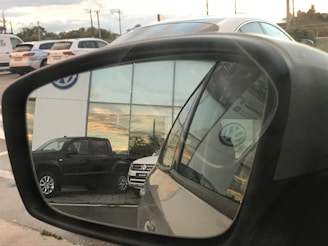 A reflection in a car side mirror shows a parking area with several vehicles, including a dark-colored truck and a light-colored SUV. The background features a building with large windows reflecting a sunset. The Volkswagen logo is visible on the building and on the vehicle registration in the mirror.