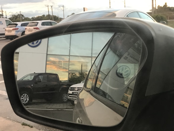 A reflection in a car side mirror shows a parking area with several vehicles, including a dark-colored truck and a light-colored SUV. The background features a building with large windows reflecting a sunset. The Volkswagen logo is visible on the building and on the vehicle registration in the mirror.