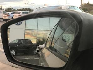 A reflection in a car side mirror shows a parking area with several vehicles, including a dark-colored truck and a light-colored SUV. The background features a building with large windows reflecting a sunset. The Volkswagen logo is visible on the building and on the vehicle registration in the mirror.