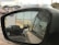 A reflection in a car side mirror shows a parking area with several vehicles, including a dark-colored truck and a light-colored SUV. The background features a building with large windows reflecting a sunset. The Volkswagen logo is visible on the building and on the vehicle registration in the mirror.