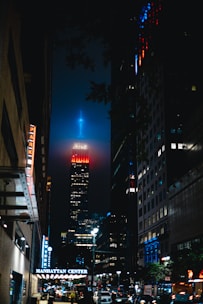 A nighttime urban scene with a towering skyscraper in the center, illuminated with vivid red and blue lights at its peak. Surrounding the main building are other tall structures, with Manhattan Center prominently visible on a sign. The street below is bustling with vehicle traffic and streetlights, casting a lively glow.