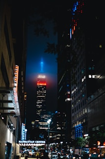 A nighttime urban scene with a towering skyscraper in the center, illuminated with vivid red and blue lights at its peak. Surrounding the main building are other tall structures, with Manhattan Center prominently visible on a sign. The street below is bustling with vehicle traffic and streetlights, casting a lively glow.