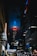 A nighttime urban scene with a towering skyscraper in the center, illuminated with vivid red and blue lights at its peak. Surrounding the main building are other tall structures, with Manhattan Center prominently visible on a sign. The street below is bustling with vehicle traffic and streetlights, casting a lively glow.