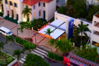 A bird's-eye view of an urban area featuring a casino with prominent neon signage. Several palm trees line the sidewalk, and a white van is parked on the street. The architecture of nearby buildings includes a mix of modern and classic designs.