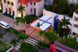 A bird's-eye view of an urban area featuring a casino with prominent neon signage. Several palm trees line the sidewalk, and a white van is parked on the street. The architecture of nearby buildings includes a mix of modern and classic designs.