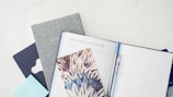A collection of stationary items including a monthly planner, a bullet journal, a decorative notebook, and a pen. The arrangement is on a light, clean surface, featuring a combination of both minimal and artistic designs.