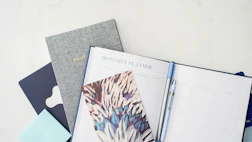A collection of stationary items including a monthly planner, a bullet journal, a decorative notebook, and a pen. The arrangement is on a light, clean surface, featuring a combination of both minimal and artistic designs.