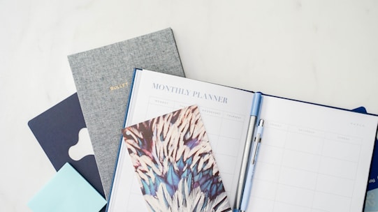 A collection of stationary items including a monthly planner, a bullet journal, a decorative notebook, and a pen. The arrangement is on a light, clean surface, featuring a combination of both minimal and artistic designs.