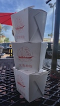 Three white Chinese takeout boxes are stacked on top of each other on an outdoor table. Each box features a red illustration of a traditional sailboat and Chinese characters. The setting appears to be outside a cafe or restaurant, with a red umbrella and palm trees visible in the background.