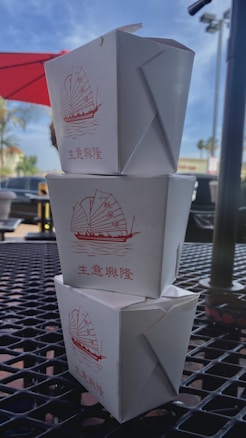 Three white Chinese takeout boxes are stacked on top of each other on an outdoor table. Each box features a red illustration of a traditional sailboat and Chinese characters. The setting appears to be outside a cafe or restaurant, with a red umbrella and palm trees visible in the background.