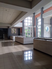 A spacious and well-lit interior space featuring large windows with a grid design, allowing natural light to flood the area. The tiled floor reflects the sunlight, creating a bright and airy atmosphere. Concrete beams and columns add a modern architectural touch, and a staircase is visible in the background, enhancing the sense of depth.
