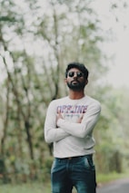 A man stands confidently in an outdoor setting with arms crossed. He is wearing sunglasses and a long-sleeve shirt with the word 'OFFLINE' printed in large letters. The background features blurred trees and greenery, suggesting a natural environment.