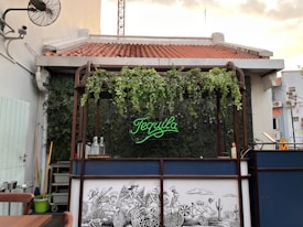 An outdoor bar features a neon sign with the word 'Tequila', surrounded by lush green foliage. The roof is tiled in red, and the bar area includes glass bottles and various items. Below the bar counter, a black and white mural depicts skeletons engaged in a humorous race. The setting is casual with nearby furniture and potted plants.