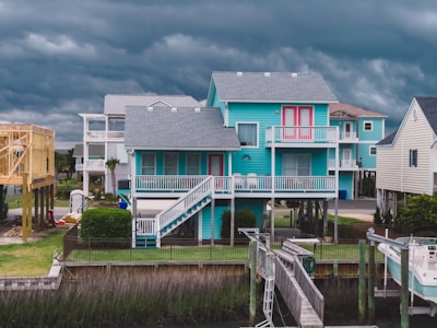 An image of a turquoise two-story house with a white wooden porch, located by a waterfront. The house has a staircase leading to the first floor and a balcony at the second floor. The sky in the background is filled with dark, stormy clouds. There are other similar houses in the vicinity and a boat moored nearby.