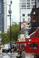 A rainy urban street scene featuring a red restaurant with a covered outdoor seating area. Pedestrians are seen holding umbrellas, and cars are parked along the street. High-rise buildings and trees line the background, and a vintage-style neon sign is visible advertising brunch. Reflections on the wet pavement add to the atmosphere.
