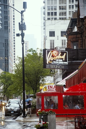 A rainy urban street scene featuring a red restaurant with a covered outdoor seating area. Pedestrians are seen holding umbrellas, and cars are parked along the street. High-rise buildings and trees line the background, and a vintage-style neon sign is visible advertising brunch. Reflections on the wet pavement add to the atmosphere.