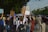 A group of people are marching on a street holding various signs and banners. One of the signs clearly reads 'Planet Over Profit', indicating a protest or demonstration focused on environmental or social issues. There are trees lining the street, and a building is visible in the background. A police officer appears to be overseeing the event.