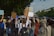 A group of people are marching on a street holding various signs and banners. One of the signs clearly reads 'Planet Over Profit', indicating a protest or demonstration focused on environmental or social issues. There are trees lining the street, and a building is visible in the background. A police officer appears to be overseeing the event.