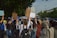 A group of people are marching on a street holding various signs and banners. One of the signs clearly reads 'Planet Over Profit', indicating a protest or demonstration focused on environmental or social issues. There are trees lining the street, and a building is visible in the background. A police officer appears to be overseeing the event.