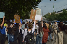 A group of people are marching on a street holding various signs and banners. One of the signs clearly reads 'Planet Over Profit', indicating a protest or demonstration focused on environmental or social issues. There are trees lining the street, and a building is visible in the background. A police officer appears to be overseeing the event.