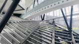 A modern architectural interior featuring large glass walls and steel beams. The structure has angular and geometric designs, with a prominent staircase or ramp intersecting the space. Natural light streams through the windows, illuminating the airy and spacious environment.