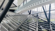 A modern architectural interior featuring large glass walls and steel beams. The structure has angular and geometric designs, with a prominent staircase or ramp intersecting the space. Natural light streams through the windows, illuminating the airy and spacious environment.