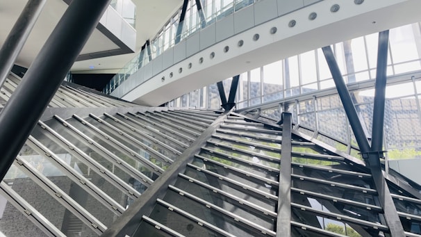 A modern architectural interior featuring large glass walls and steel beams. The structure has angular and geometric designs, with a prominent staircase or ramp intersecting the space. Natural light streams through the windows, illuminating the airy and spacious environment.