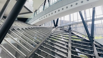 A modern architectural interior featuring large glass walls and steel beams. The structure has angular and geometric designs, with a prominent staircase or ramp intersecting the space. Natural light streams through the windows, illuminating the airy and spacious environment.