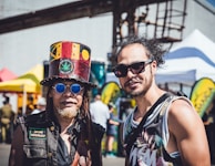 Two men are outdoors at a festival or market. The man on the left wears a colorful top hat featuring Bob Marley insignia and blue round sunglasses. He has a beard and wears a vest with patches. The man on the right wears sunglasses and a sleeveless printed top. Brightly colored tents and a sunny sky can be seen in the background.