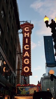 A large illuminated marquee sign displaying the word 'Chicago' vertically on the side of a theater. The sign is adorned with bright lights and is situated among tall buildings. Below the marquee, a colorful advertisement for a show is visible, while a street lamp glows against a backdrop of a clear evening sky.