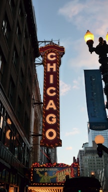A large illuminated marquee sign displaying the word 'Chicago' vertically on the side of a theater. The sign is adorned with bright lights and is situated among tall buildings. Below the marquee, a colorful advertisement for a show is visible, while a street lamp glows against a backdrop of a clear evening sky.