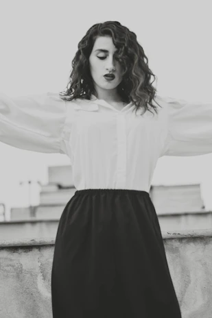 A model wearing a flowing white dress with subtle black patterns, standing in a sunlit urban street.