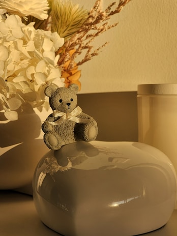 A small teddy bear with a bow sits atop a smooth, glossy surface, accompanied by a serene arrangement of dried flowers and foliage in muted tones.