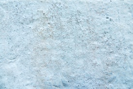 A textured surface that appears to be a close-up of a wall with a rough, uneven finish. The surface is primarily a light blue color with areas of darker speckles and subtle variations.