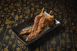 On a dark plate, three crispy fried chicken wings are artfully arranged. The background is covered in an intricate pattern of elegant gold calligraphy on a textured black surface, creating a sophisticated atmosphere.