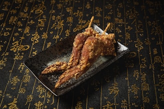 On a dark plate, three crispy fried chicken wings are artfully arranged. The background is covered in an intricate pattern of elegant gold calligraphy on a textured black surface, creating a sophisticated atmosphere.