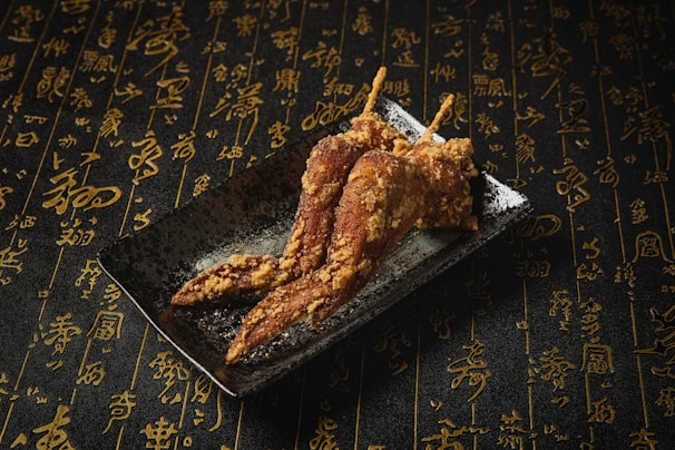On a dark plate, three crispy fried chicken wings are artfully arranged. The background is covered in an intricate pattern of elegant gold calligraphy on a textured black surface, creating a sophisticated atmosphere.